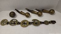 Overview photo showing three brass hanging hook-shaped pieces, a large brass bell, and a leather strap with five attached brass horse brasses.