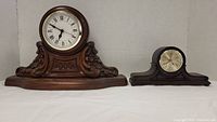 Both vintage wood carved mantle clocks displayed side by side on white background surface.