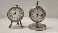 Photo showing both metal clocks side by side, one pocket watch style on tripod and one round on circular base, front view.
