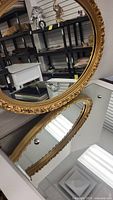 Photo showing the oval gilt framed mirror with reflected store shelving and other decorative items in the background. Highlights the ornate details of the frame.
