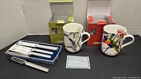 Two ceramic mugs in boxes with bird illustrations and two sets of Rada stainless steel cutting utensils in boxes, plus a peeler, all displayed on a black surface.