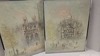 Two framed paintings showing Paris landmarks Notre-Dame Cathedral and Sacré-Cœur Basilica in soft muted tones with delicate people and tree elements.