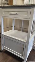 Front view of white cabinet showing granite top, upper drawer, middle shelf, lower cabinet door, and caster wheels