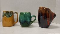 Three ceramic mugs displayed together: yellow Beauce mug, green mug, brown textured mug.