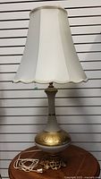 Full view of vintage table lamp with white shade and gold and white decorative base on wooden table against slat wall