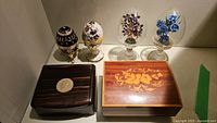 Group shot showing two bejeweled egg music boxes, two crystal eggs under glass domes, two wooden boxes