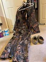 Photo showing men's XL camouflage flannel hunting jacket on hanger, matching pants laid out, and pair of camo Crocs shoes on floor.