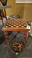 Full view of chess table showing wooden inlaid chessboard on top, carved legs, and basket of chess pieces underneath