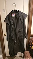 Full view of the black oilskin duster coat hanging on a door, showing length and button closure.