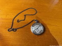 Full view of the Tawek Präzision pocket watch with chain laid out on a wooden table surface. Shows watch front and attached chain clearly.
