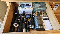 Overview of drawer showing boxed Night Beam lantern, boxed trail camera, unboxed camouflage trail camera, white jump starter and multiple flashlights