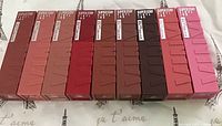 Front view of nine Maybelline New York Superstay Vinyl Ink liquid lipsticks in their original boxes, arranged showing the range of colours.
