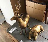 Pair of solid metal deer figurines with brass-colored finish, different sizes, standing on a wooden surface.