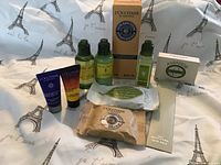 All L’Occitane beauty products displayed on Eiffel Tower print fabric, showing full size hand balm in box and various travel size bottles, tubes, wrapped soaps, and nail files.