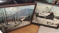View of both framed winter landscape paintings leaning against a wall showing overall size and framing