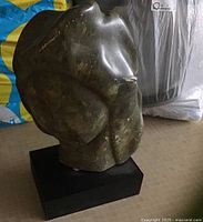 Stone carved sculpture on black rectangular base, showing front and right side view. Figure has a dark greenish tone with lighter veining and scratches visible on surface.