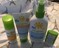Four sealed Babyganics sunscreen products: lotion, spray, two sticks including Kids by Babyganics tropical stick. Displayed on fabric with Eiffel Tower print.