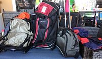Group photo of five travel bags including a large silver and black hiking backpack, a black and red wheeled backpack, a compact gray roller bag, and two other bags piled together on a blue floor.