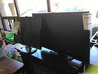 Photo showing two black computer monitors on a black desk, one larger than the other, lit by daylight from a window.