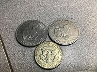 Two 1972 one dollar coins and one 1966 half dollar coin shown from the reverse side featuring eagle designs and inscriptions.