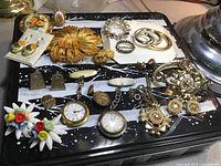 Photo showing a variety of vintage costume jewellery. Includes floral ceramic earrings and brooch, large gold-tone swirl brooch, rhinestone brooches, cufflinks, nurse watches, and pins scattered on a black and white surface.