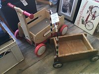 Two vintage wooden children’s push toys including a Mobo Baby Walker with wooden body and red wheels and handles, and a smaller Hape wooden toy wagon with painted handle and rubber wheels.