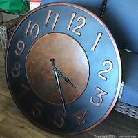 Photo showing the large round clock leaning against a sofa, highlighting the black and bronze color scheme and raised numerals.