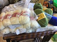 Image showing 24 balls of natural white White Rose Sayelle yarn in plastic bag and 1 roll blue and 2.5 balls green acrylic yarn on table.
