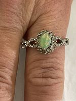 Ring worn on finger showing oval synthetic opal center stone with surrounding clear cubic zirconia stones in a detailed sterling silver setting