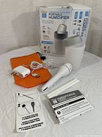 Humidifier device with power adapter, USB cable, two white sticks, user manuals, and orange carrying pouch all displayed in front of the product box.