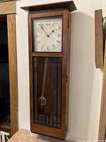 Full view of the Ethan Allen wall clock mounted on the wall showing the rectangular wooden body, clock face, and pendulum behind decorative glass.