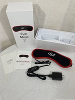 Full contents laid out: DPL branded box, black and red eye mask, black power adapter, user and safety manuals.