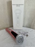 Pink handheld facial rejuvenation device next to white USB charging cable and white branded box with product image and title