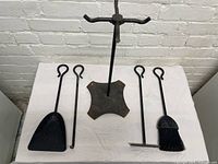 Photo of the complete fireplace tool set including a metal stand with cross-shaped base and four tools (shovel, tongs, poker, brush) on a white cloth surface in front of a white brick wall.