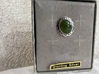 Front view of the jade and sterling silver tie pin with visible green jade cabochon set in a decorative sterling silver bezel, placed on a gray fabric box marked 'Sterling Silver'.