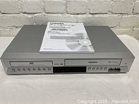 Front view of Toshiba SD-V383SC dual DVD and VHS player recorder showing device with multiple control buttons, disc and cassette slots, and owner's manual placed on top.
