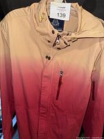 Front view of American Rag ombré anorak jacket hanging, showing zipper and snap button closure, long sleeves, and gradient from cream to red.