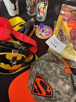 Batman ceramic mug with yellow logo on black background, Thor figure, red and yellow Wonder Woman apron fabric, birthday button, and some drinkware on black cloth.