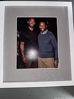 Photo showing a framed photo of two men, one in black shirt and the other in grey sweater, with neutral tone mat and white frame.