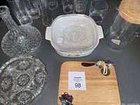 Full view of all items including wooden cheese board with turtle spreader knife, cut glass decanter and ashtray, Corning Ware casserole dish, glass jar with cork lid, and 4 clear glasses.