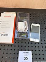 Photo showing iPhone SE phone powered on with the start-up screen, accessories including charger, USB cable and earphones in plastic tray, alongside certified smartphone box.