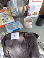 KitchenAid stainless steel mixing bowl, citrus juicer box, ceramic mugs, metal pot rack, and kitchen utensils on table.
