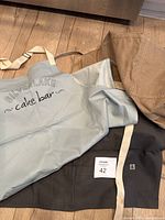 Light gray Silverlake Cake Bar apron showcased on top of black and brown Chef Works charcoal bistro aprons showing branding and fabric material.