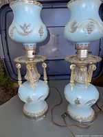 Pair of vintage blue glass lamps shown together from different angles, displaying their blue opaque glass with floral and grapevine designs, metal fittings, and faux crystal drop decorations. Electrical cords visible, lamps set on grey surface with dark blue background.