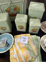 Vintage bread box and four repainted canisters with floral design, Norman Rockwell plates, and folded dish towels arranged on black surface.