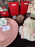 Red MasterWare metal canisters labeled sugar, flour, and coffee, large pink Lu-Ray pastel platter, pink glass reamer with ridged top, and vintage translucent white apron with red trim, all arranged together