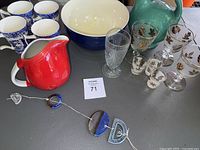 Overview of all items including red Hall pitcher, Metlox green pitcher, blue and white Spode mugs, large bowl, various stemmed and shot glasses, and decorative ceramic hanging piece.