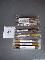 Photo showing eight wooden ballpoint pens neatly arranged on a flat surface. Each pen is individually packaged in clear plastic tubes with white labels describing wood type and metal trim. Pens include walnut, wenge, maple crotch, zebra wood, fiddleback maple, maple burle, and olivewood.