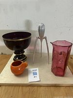 Full view of four items: orange Metlox creamer, deep ruby red pedestal bowl with gold trim, silver tripod aluminum citrus reamer by Alessi, and pink faceted studio art glass carafe.