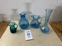 Wide view of four vintage glass pieces on a wooden shelf showing two crackle glass pitchers, one crackle glass vase, and one tall narrow-neck jug